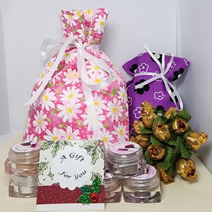 May include: A pink fabric drawstring bag with a white daisy pattern, a purple fabric drawstring bag with a black and white flower pattern, a small bouquet of gold-colored artificial roses, and several clear cosmetic jars with a gift card reading A Gift For You.