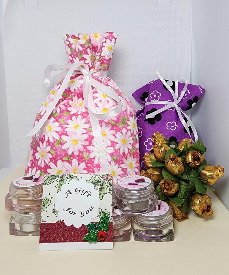 May include: A pink fabric drawstring bag with a white daisy pattern, a purple fabric drawstring bag with a black and white flower pattern, a small bouquet of gold-colored artificial roses, and several clear cosmetic jars with a gift card reading A Gift For You.