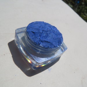 May include: A small, clear container filled with a bright blue glitter eyeshadow. The container is made of plastic and has a clear lid.