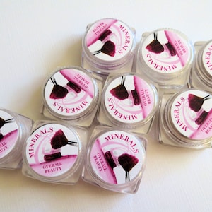 May include: Eight small, clear plastic cosmetic jars with screw-on lids. Each lid features a circular white label with pink accents, a graphic of a makeup brush, and the printed text "MINERALS" and "OVERALL BEAUTY" arranged in a circular pattern.