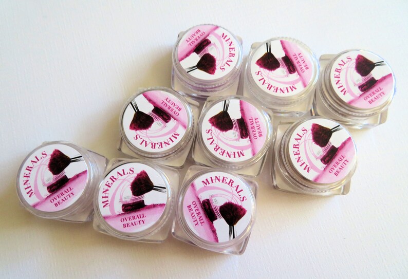 May include: Eight small, clear plastic cosmetic jars with screw-on lids. Each lid features a circular white label with pink accents, a graphic of a makeup brush, and the printed text "MINERALS" and "OVERALL BEAUTY" arranged in a circular pattern.