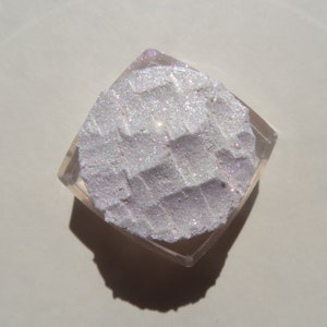 Op de afbeelding: Een vierkant, helder cosmetica-compact met een getextureerd, glinsterend, lichtpaars poeder. Het poeder heeft een rasterpatroon en is omgeven door een heldere plastic behuizing.