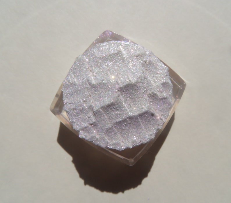 Op de afbeelding: Een vierkant, helder cosmetica-compact met een getextureerd, glinsterend, lichtpaars poeder. Het poeder heeft een rasterpatroon en is omgeven door een heldere plastic behuizing.