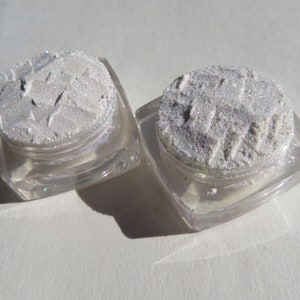 May include: Two small, square, clear plastic cosmetic jars filled with a shimmering, silver-toned powder. The powder has a textured, pressed surface with visible indentations and a reflective, metallic finish that catches the light.