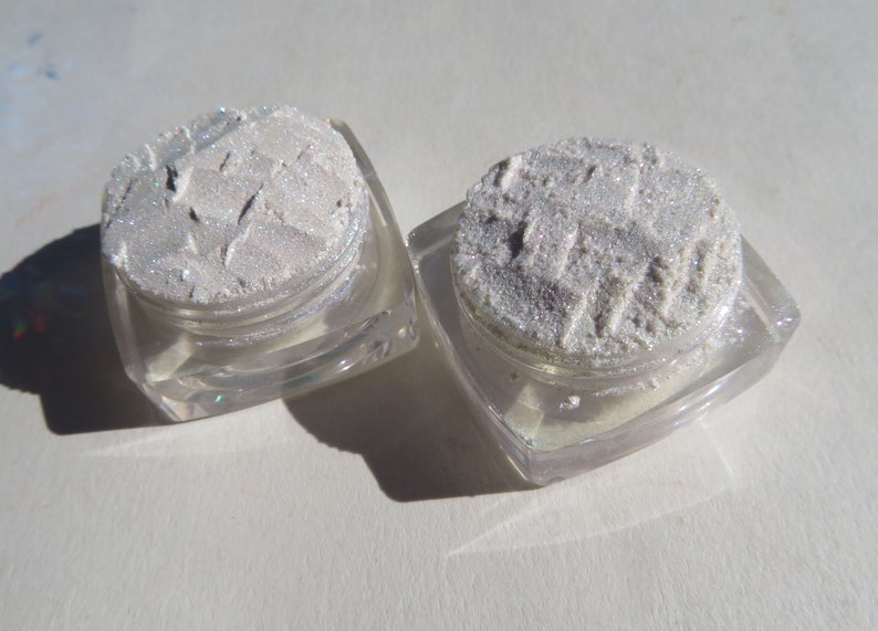 May include: Two small, square, clear plastic cosmetic jars filled with a shimmering, silver-toned powder. The powder has a textured, pressed surface with visible indentations and a reflective, metallic finish that catches the light.