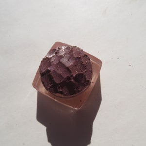 May include: A small, square container with a clear lid, filled with a deep purple eyeshadow with a shimmery finish.