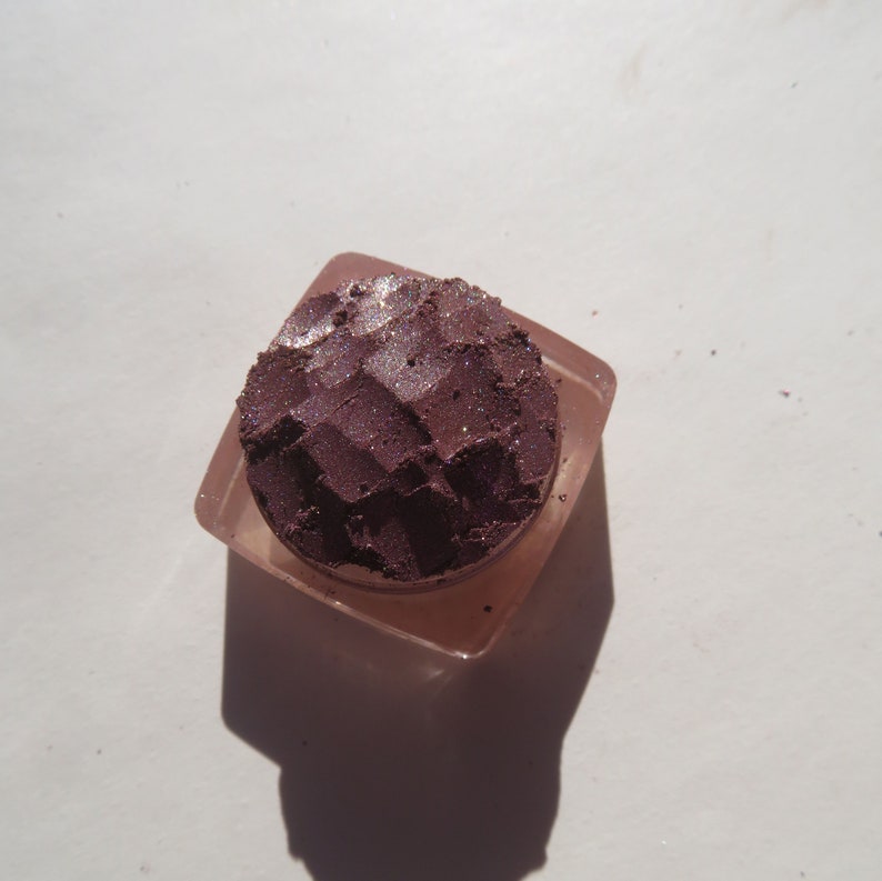 May include: A small, square container with a clear lid, filled with a deep purple eyeshadow with a shimmery finish.