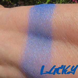May include: A close-up of a light blue eyeshadow with a shimmery finish. The eyeshadow is swatched on a person's arm. The word "Lucky" is written in blue letters below the swatch.