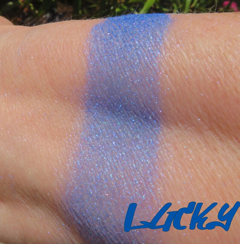 May include: A close-up of a light blue eyeshadow with a shimmery finish. The eyeshadow is swatched on a person's arm. The word "Lucky" is written in blue letters below the swatch.