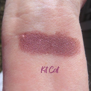 May include: A close-up of a shimmery, mauve-toned eyeshadow with a fine glitter finish. The eyeshadow is swatched on a person's arm and labeled "Kit Cat".