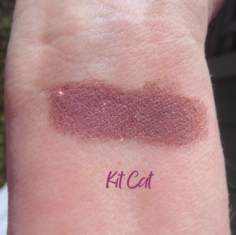 May include: A close-up of a shimmery, mauve-toned eyeshadow with a fine glitter finish. The eyeshadow is swatched on a person's arm and labeled "Kit Cat".