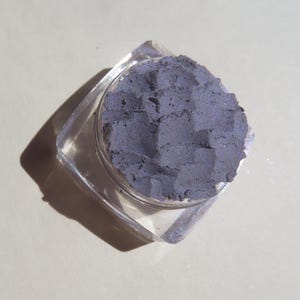 May include: A small, clear plastic container with a lid, filled with a dark gray eyeshadow. The eyeshadow has a slightly textured surface.