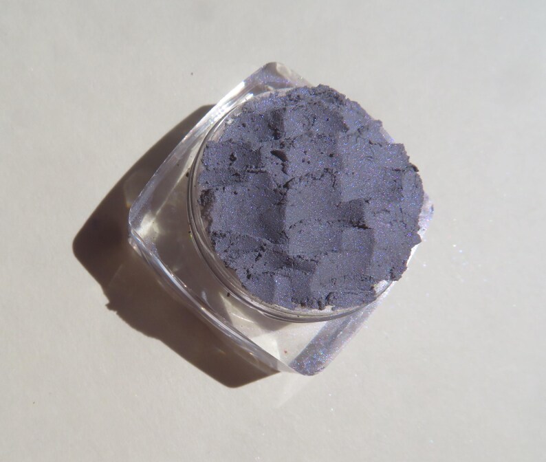 May include: A small, clear plastic container with a lid, filled with a dark gray eyeshadow. The eyeshadow has a slightly textured surface.