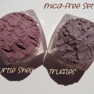May include: Two small containers of mica-free eyeshadow. The eyeshadow on the left is a light purple shade labelled "Turtle Shell". The eyeshadow on the right is a dark grey shade labelled "Truffles".