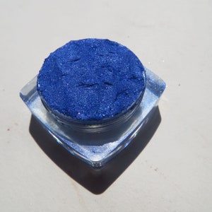 May include: A small, round container filled with a deep blue glitter eyeshadow. The container is clear and has a silver lid.