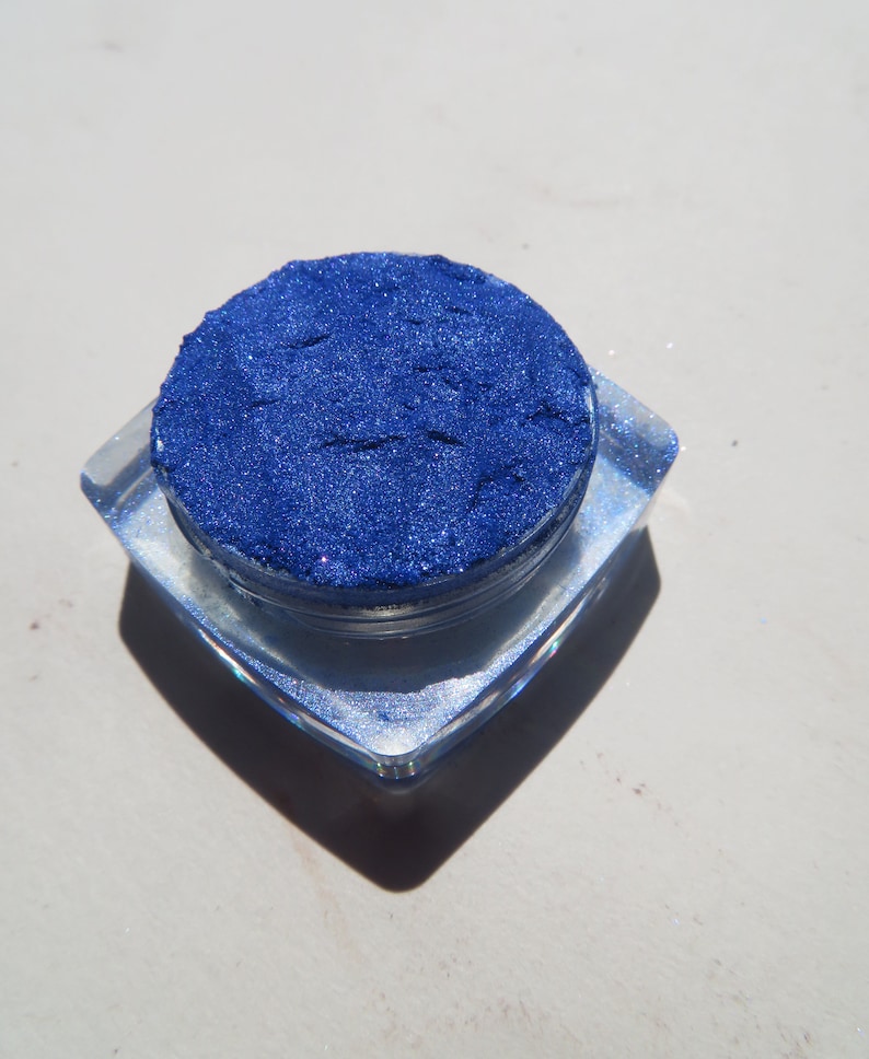 May include: A small, round container filled with a deep blue glitter eyeshadow. The container is clear and has a silver lid.