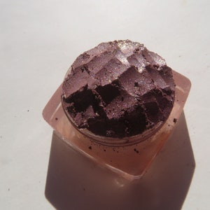 May include: A small, round container of loose, shimmery purple eyeshadow with a textured surface. The eyeshadow is in a clear plastic container with a pink lid.