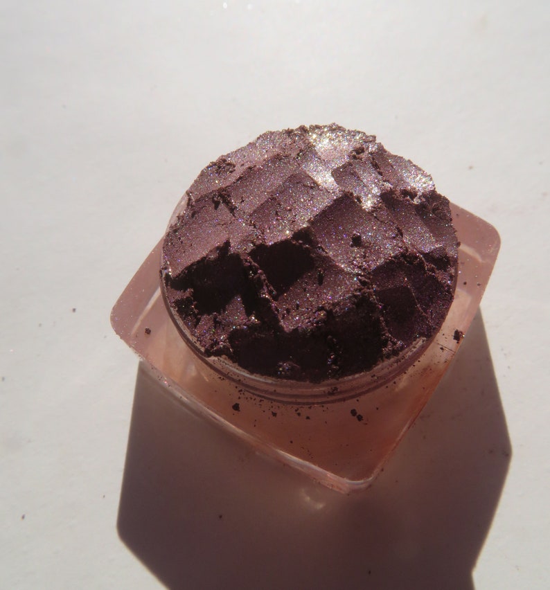 May include: A small, round container of loose, shimmery purple eyeshadow with a textured surface. The eyeshadow is in a clear plastic container with a pink lid.