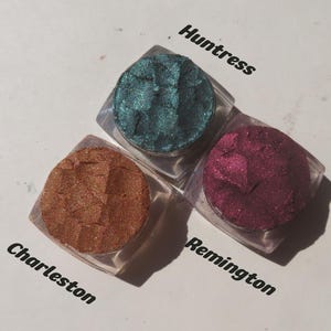 May include: Three square clear containers of loose powder eyeshadow in metallic finishes. The colors are a deep teal labeled Huntress, a warm copper labeled Charleston, and a rich magenta labeled Remington. Each pigment has a textured surface.