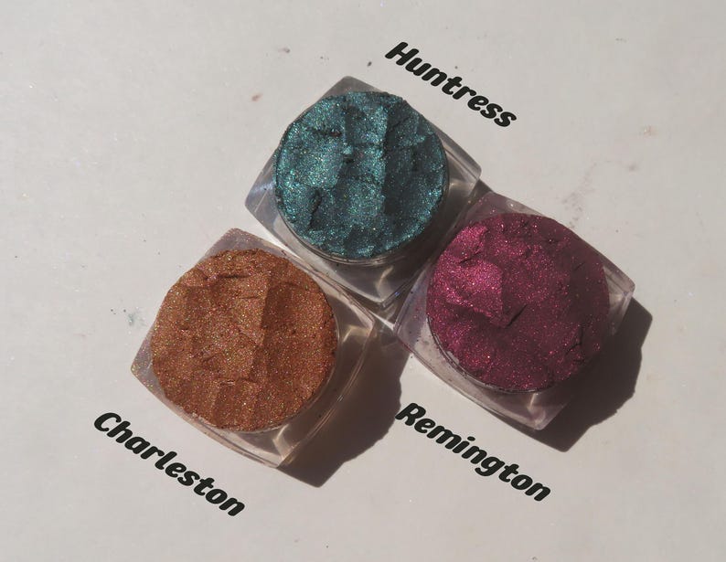 May include: Three square clear containers of loose powder eyeshadow in metallic finishes. The colors are a deep teal labeled Huntress, a warm copper labeled Charleston, and a rich magenta labeled Remington. Each pigment has a textured surface.
