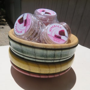 May include: A stack of three round wooden bowls in shades of green, yellow, and pink, containing several clear plastic jars of Minerals Overall Beauty cosmetic powder, each featuring a label with a graphic of a makeup brush and pink accents.
