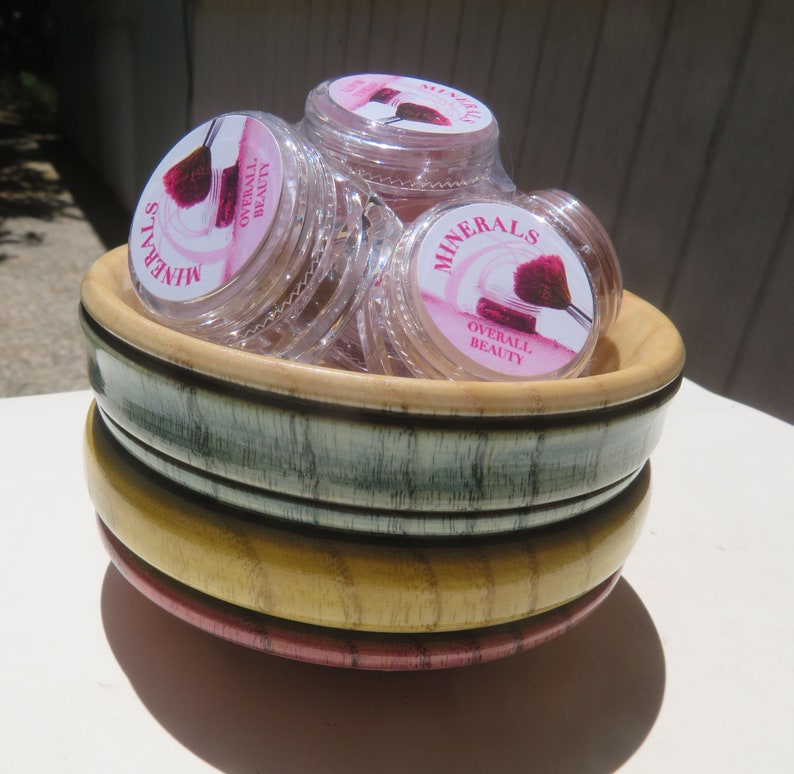 May include: A stack of three round wooden bowls in shades of green, yellow, and pink, containing several clear plastic jars of Minerals Overall Beauty cosmetic powder, each featuring a label with a graphic of a makeup brush and pink accents.
