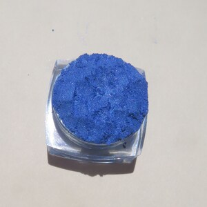 May include: A small round container of blue eyeshadow. The eyeshadow is a shimmery blue color.