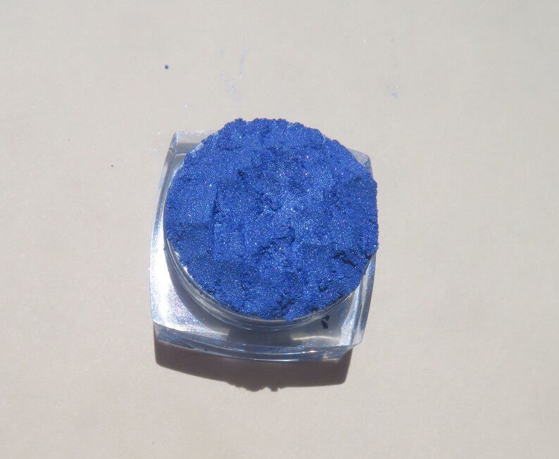 May include: A small round container of blue eyeshadow. The eyeshadow is a shimmery blue color.