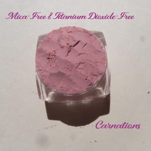 May include: A small, clear, square cosmetic jar filled with dusty rose-colored powder. The text "Mica-Free & Titanium Dioxide-Free" appears at the top, and the word "Carnations" is printed in the bottom right corner in a purple script font.