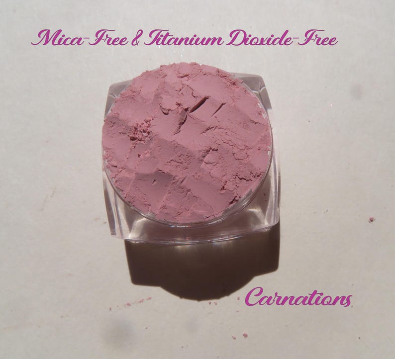 May include: A small, clear, square cosmetic jar filled with dusty rose-colored powder. The text "Mica-Free & Titanium Dioxide-Free" appears at the top, and the word "Carnations" is printed in the bottom right corner in a purple script font.