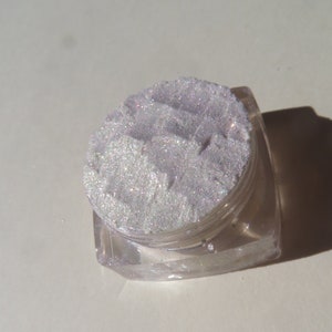 Op de afbeelding: Een kleine, heldere, vierkante pot gevuld met glinsterend, lichtpaars cosmetisch poeder. Het poeder heeft een textuur en lijkt fijn gemalen. De pot staat op een witte ondergrond.