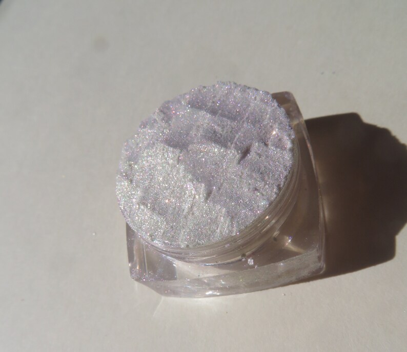 Op de afbeelding: Een kleine, heldere, vierkante pot gevuld met glinsterend, lichtpaars cosmetisch poeder. Het poeder heeft een textuur en lijkt fijn gemalen. De pot staat op een witte ondergrond.