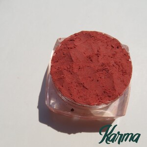Op de afbeelding: Een kleine, ronde verpakking met rood poedermake-up. De verpakking is van doorzichtig plastic en heeft een label met de tekst "Karma".