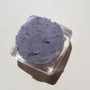 May include: A small, clear plastic container with a lid, filled with a dark gray eyeshadow. The eyeshadow has a slightly shimmery finish.