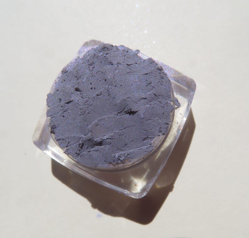 May include: A small, clear plastic container with a lid, filled with a dark gray eyeshadow. The eyeshadow has a slightly shimmery finish.