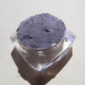 May include: A small, clear, square container with a silver lid filled with a dark gray powder. The powder has a slight shimmer.