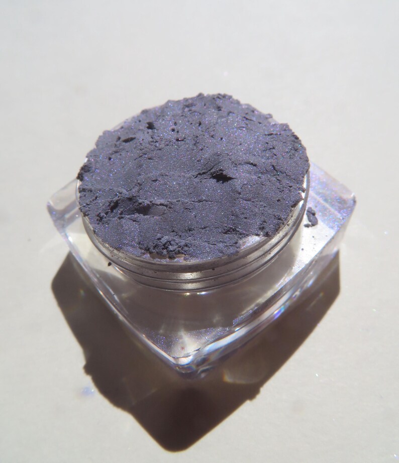 May include: A small, clear, square container with a silver lid filled with a dark gray powder. The powder has a slight shimmer.