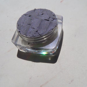May include: A small, clear container with a silver lid holds a sample of dark gray eyeshadow. The eyeshadow has a shimmery finish.