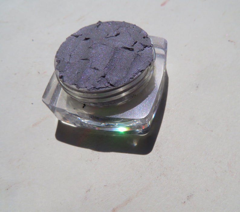 May include: A small, clear container with a silver lid holds a sample of dark gray eyeshadow. The eyeshadow has a shimmery finish.