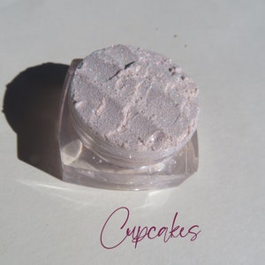 May include: A small, clear, square-based cosmetic jar filled with a pale pink, shimmery, loose powder pigment. The surface of the powder is textured and reflective. The word Cupcakes is written in a dark magenta script font below the container.