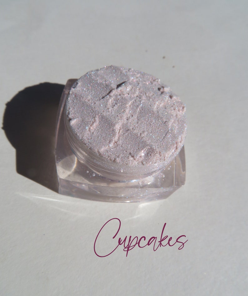May include: A small, clear, square-based cosmetic jar filled with a pale pink, shimmery, loose powder pigment. The surface of the powder is textured and reflective. The word Cupcakes is written in a dark magenta script font below the container.