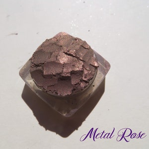 May include: A small container of loose, shimmery, metallic rose-colored eyeshadow. The eyeshadow is in a clear, square container with rounded edges.