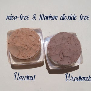 Peut inclure: Deux petits contenants ronds de poudre de maquillage min&eacute;rale en vrac. Le contenant de gauche est &eacute;tiquet&eacute; "Hazelnut" et le contenant de droite est &eacute;tiquet&eacute; "Woodlands". Le texte "mica-free & titanium dioxide free" est &eacute;crit au-dessus des contenants.