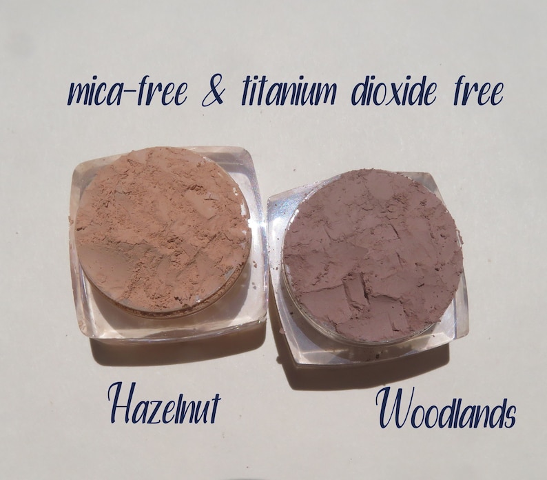 Peut inclure: Deux petits contenants ronds de poudre de maquillage min&eacute;rale en vrac. Le contenant de gauche est &eacute;tiquet&eacute; "Hazelnut" et le contenant de droite est &eacute;tiquet&eacute; "Woodlands". Le texte "mica-free & titanium dioxide free" est &eacute;crit au-dessus des contenants.