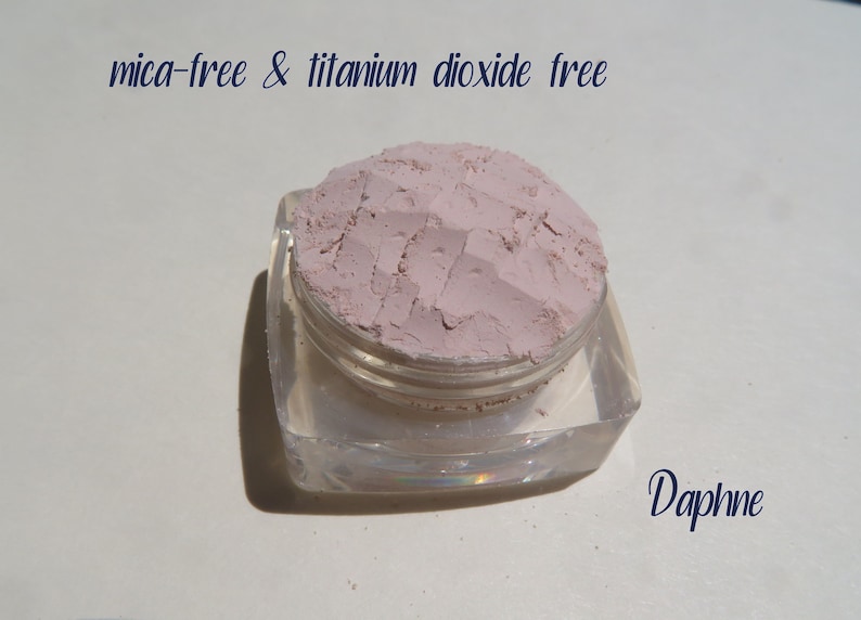 Peut inclure: Un petit r&eacute;cipient carr&eacute; transparent avec un couvercle, rempli d'une poudre rose clair. Le texte "mica-free & titanium dioxide free" est &eacute;crit au-dessus du r&eacute;cipient. Le mot "Daphne" est &eacute;crit sous le r&eacute;cipient.