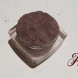 May include: A clear, square-shaped cosmetic jar filled with a dark, matte, brownish-purple powder. The surface of the powder shows visible texture and indentations. The word "Figs" is printed in a dark, cursive script to the lower right.