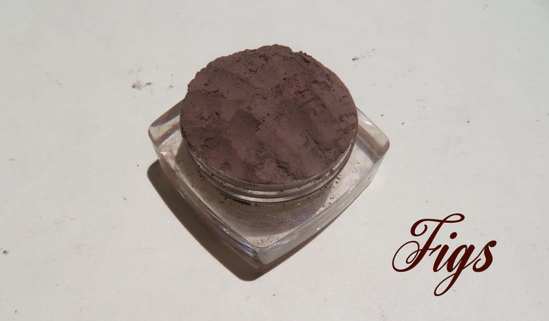 May include: A clear, square-shaped cosmetic jar filled with a dark, matte, brownish-purple powder. The surface of the powder shows visible texture and indentations. The word "Figs" is printed in a dark, cursive script to the lower right.