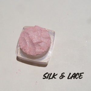 Op de afbeelding: Een klein, doorzichtig bakje gevuld met een roze en wit glinsterend poeder. De tekst "SILK & LACE" is onder het bakje gedrukt.