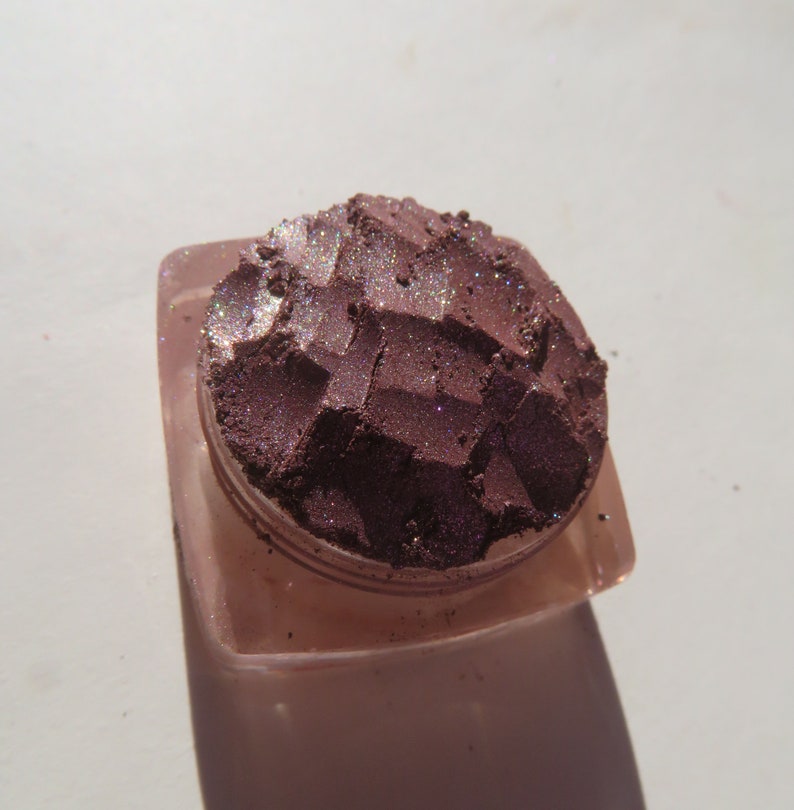 May include: A small, round container of loose, shimmery, dark purple eyeshadow with a textured surface.