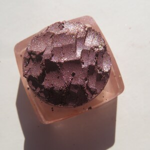 May include: A close-up of a small, round container of purple eyeshadow with a shimmery finish. The eyeshadow is in a clear container with a pink lid.